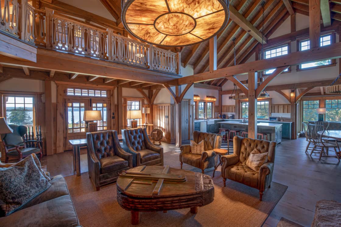 Rustic living room with high vaulted ceilings and exposed wooden beams evokes the charm of a luxury home inspired by a classic bank barn. Leather armchairs, a wooden coffee table, and large windows fill the space with natural light.