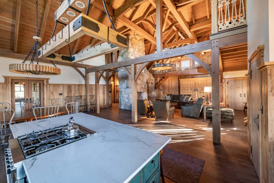 Spacious rustic kitchen and living area in a renovated bank barn, featuring exposed wooden beams, stone fireplace, marble island with stovetop, wooden chairs, and a cozy seating area with armchairs under warm natural light.