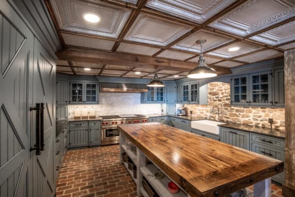 A rustic kitchen in this luxury home features a large wooden island, brick flooring, green cabinets, a farmhouse sink, stone backsplash, stainless appliances, and a coffered ceiling with recessed lighting and pendant lamps.