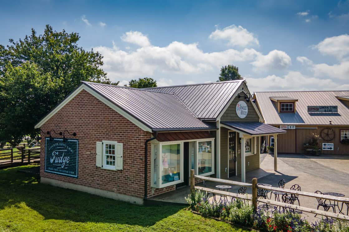A small brick and wood building with a sign reading The Fudge Shop, outdoor tables and chairs, and a chalkboard fudge menu, set on a sunny day—reminiscent of a cozy party barn amid blue sky and green grass.