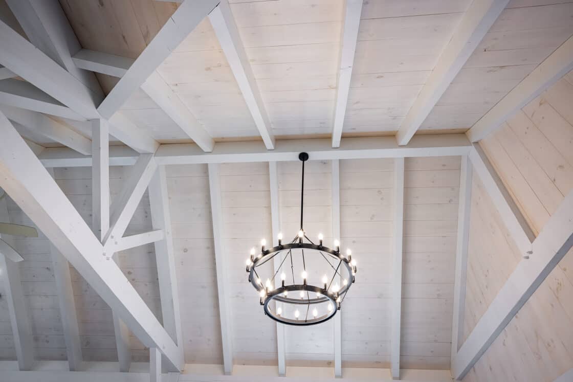 A circular black metal chandelier with lit candles hangs from a vaulted ceiling made of white-painted wooden beams and panels, adding elegance to this luxury home inspired by classic bank barn architecture.
