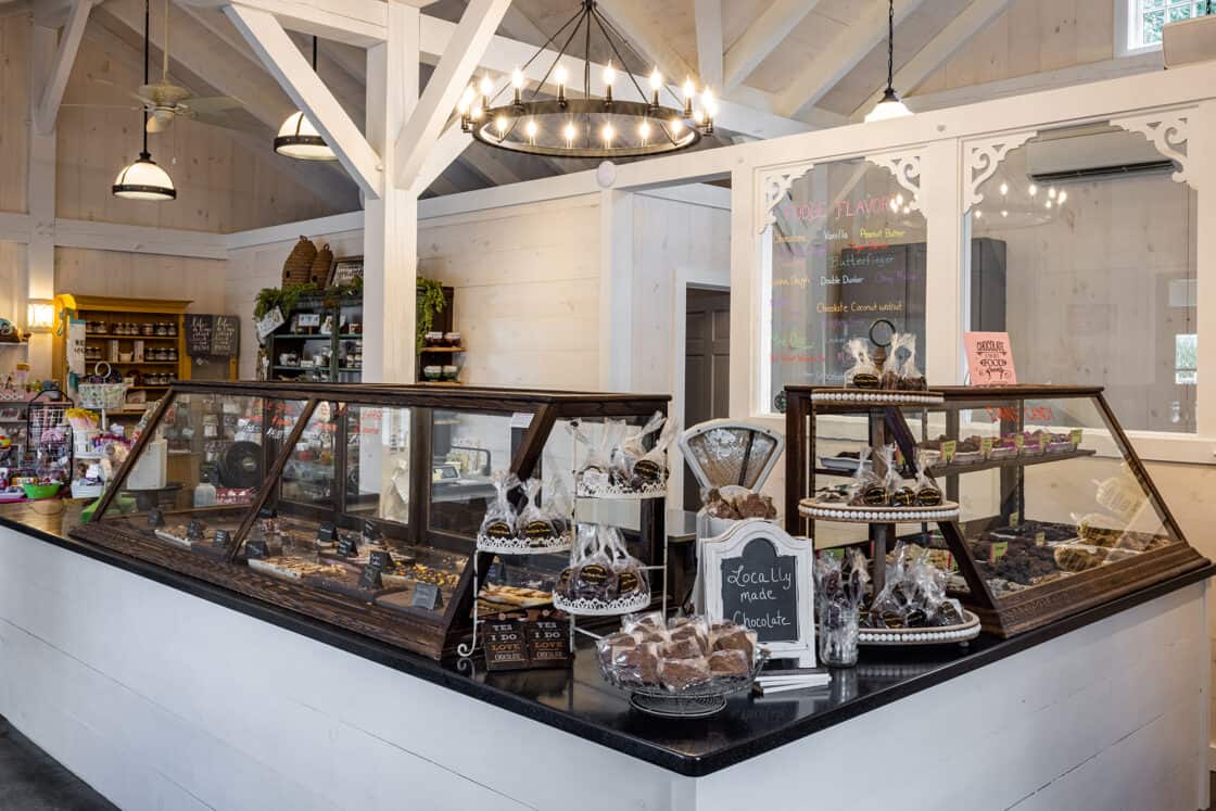 A cozy candy shop interior with white walls and wooden beams, reminiscent of a luxury horse barn, features glass display cases filled with assorted chocolates. Shelves in the back hold jars, bottles, and gift items, while a chalkboard menu completes the look.