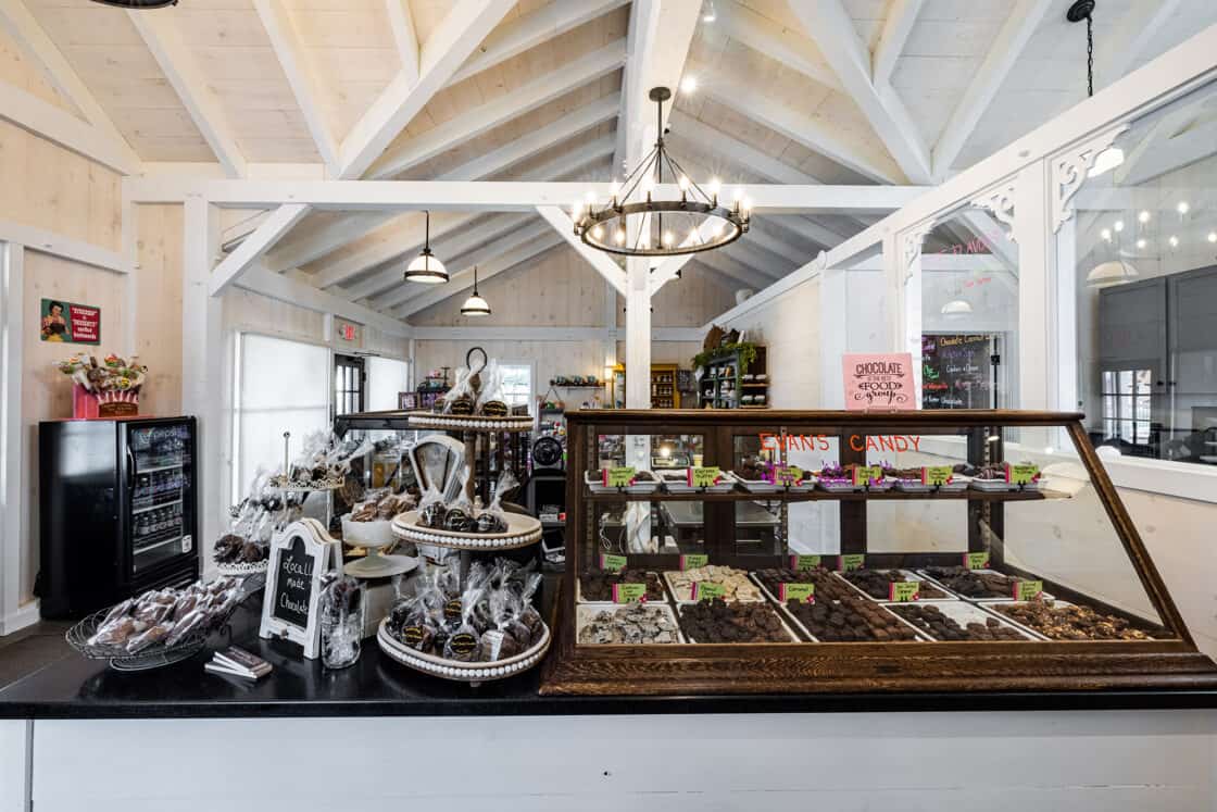 A cozy candy shop with white wooden beams reminiscent of a party barn, a glass display case filled with assorted chocolates, shelves of sweets, a tiered tray of wrapped treats, and warm lighting from chandeliers and hanging lamps.