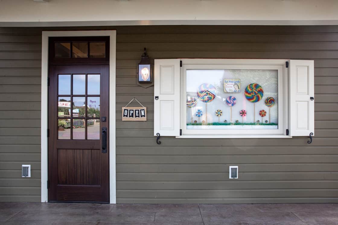 A storefront with a dark wooden door and a hanging “OPEN” sign. Next to the door is a window display of colorful lollipops and candies, framed by white shutters on a greenish-grey paneled wall—reminiscent of a charming party barn facade.
