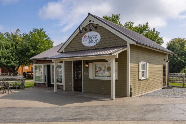 A small, tan Sweet Shop with white trim and a sign above the entrance sits on a paved area, reminiscent of a charming party barn, surrounded by trees and outdoor seating under a partly cloudy sky.