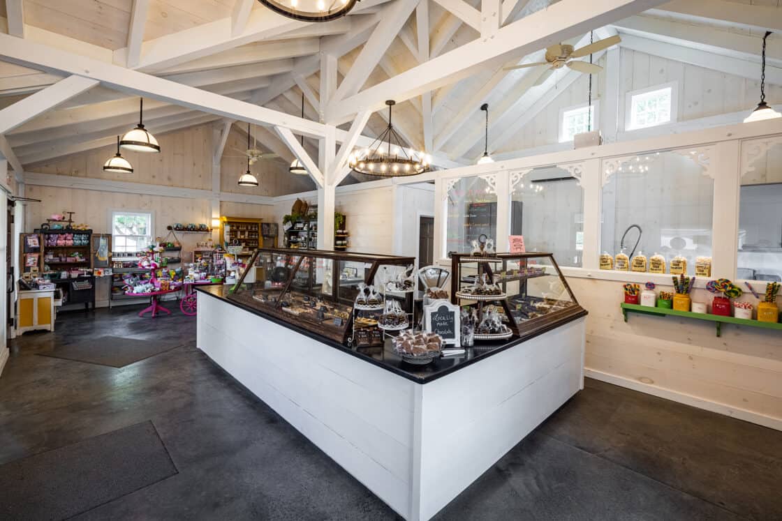 A spacious candy store with white wooden beams reminiscent of a luxury horse barn, large windows, and displays of chocolates and sweets in glass cases. Colorful candy lines the shelves, creating a bright and inviting atmosphere.