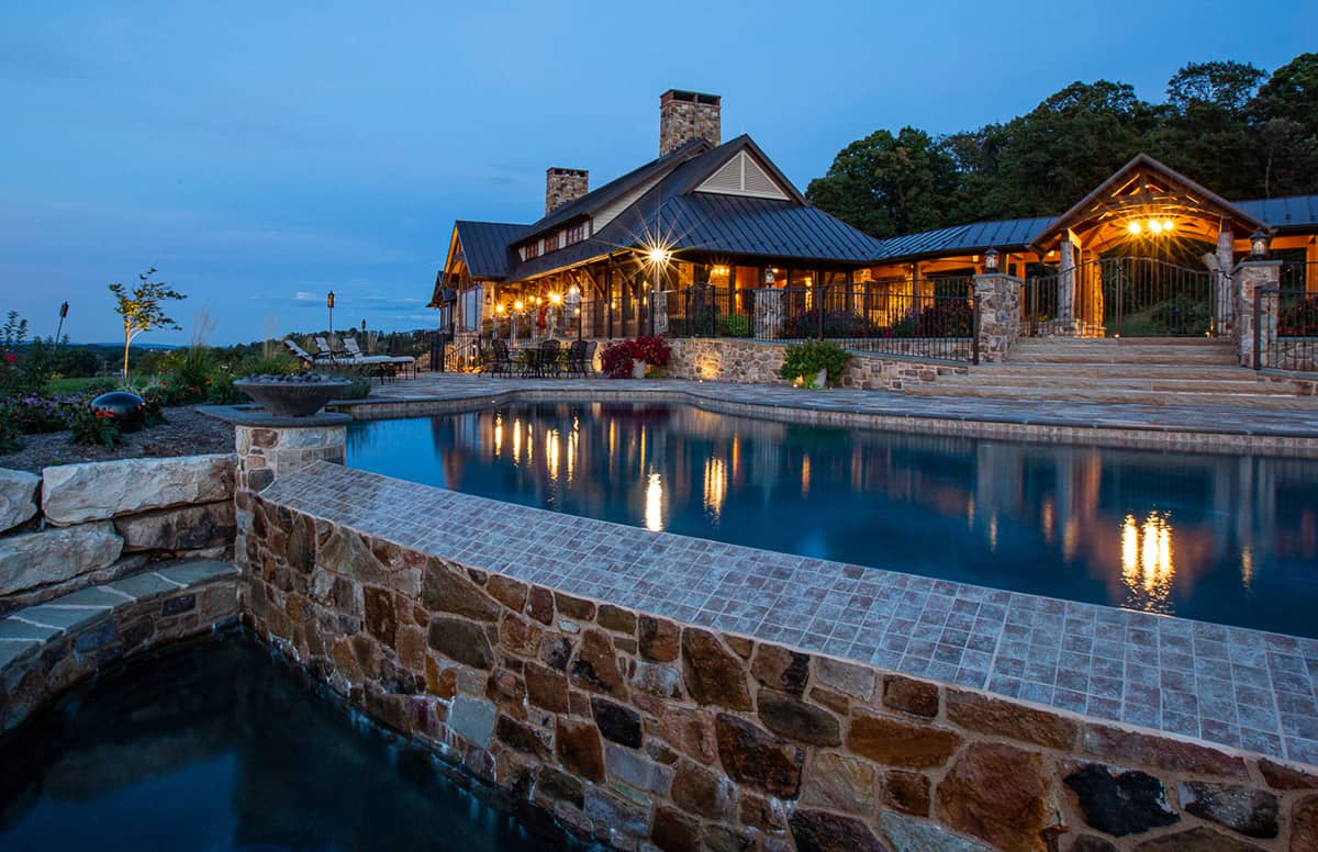 A luxurious rustic house with stone and wood exterior sits behind a lit infinity-edge pool at dusk, evoking the charm of a luxury home. Warm lights illuminate the porch and outdoor space, surrounded by landscaped gardens and trees.