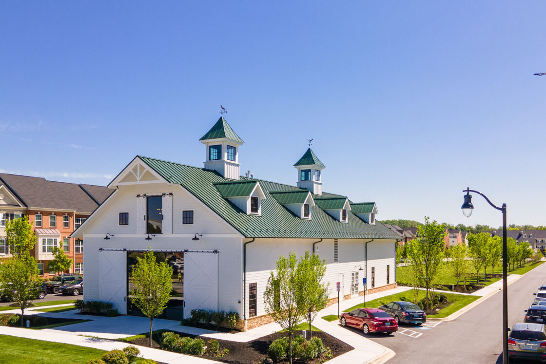A large white bank barn with a green roof and cupolas sits near a street lined with parked cars and young trees. Apartment buildings are nearby, and a plane flies in the clear blue sky above.