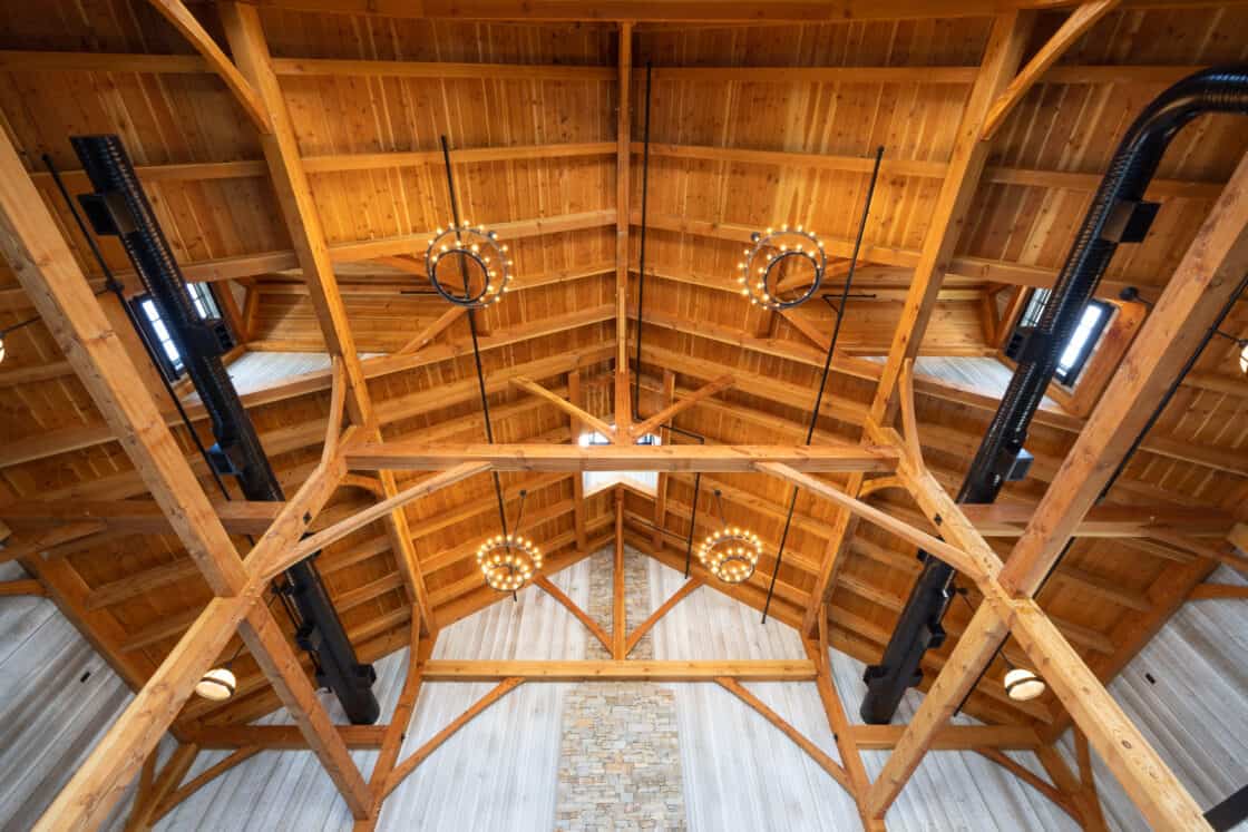 Wooden ceiling with exposed beams, circular modern chandeliers, black metal pipes, and a stone accent wall at the center create a luxury horse barn vibe. Natural light enters through small windows in the roof.