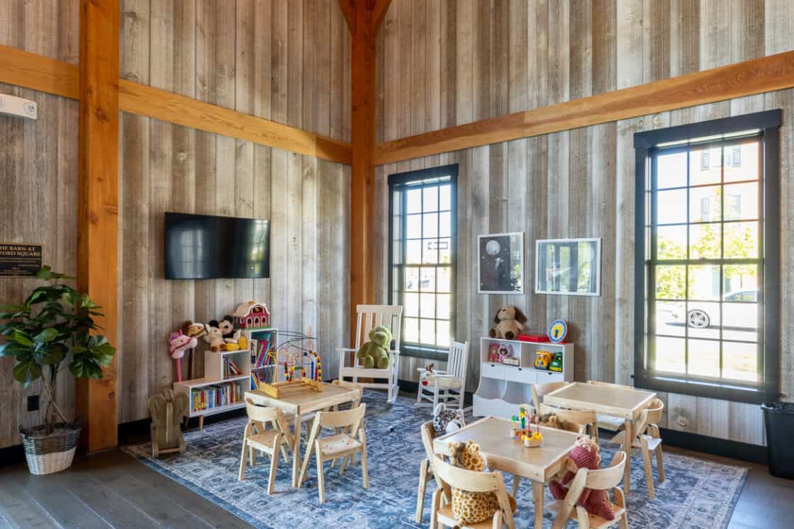 A cozy, high-ceilinged playroom in a converted bank barn features wood-paneled walls, a large rug, small tables and chairs, toys, plush animals, a TV, and big windows letting in natural light.