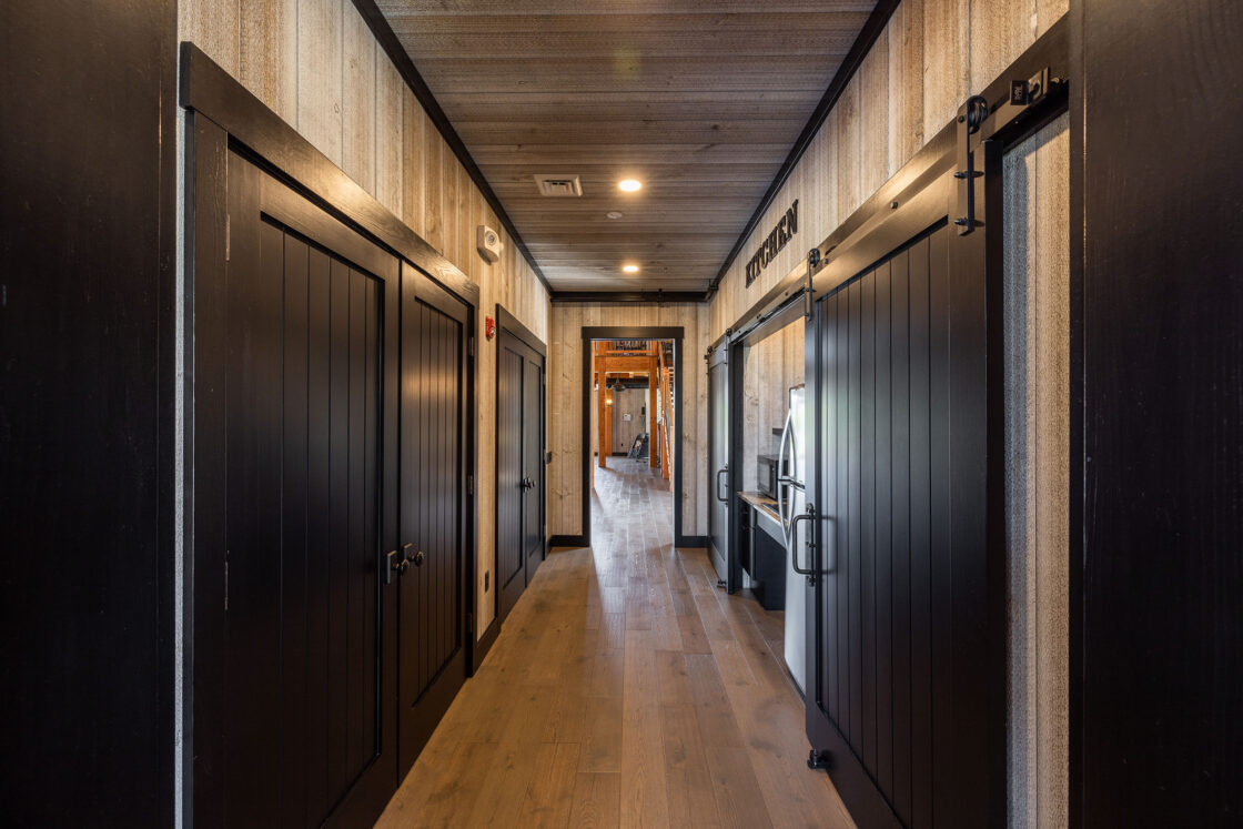 A hallway with wooden floors and black doors on both sides leads to a lit room at the end. The walls feature light-colored wood panel texture, and a sliding barn door inspired by luxury home design is visible on the right.
