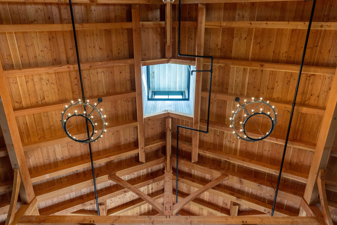 Wooden ceiling with exposed beams, two circular chandeliers glowing warmly, and a central skylight letting in natural light—perfect for a luxury horse barn or stylish party barn. Black pipes run along the ceiling toward the skylight.