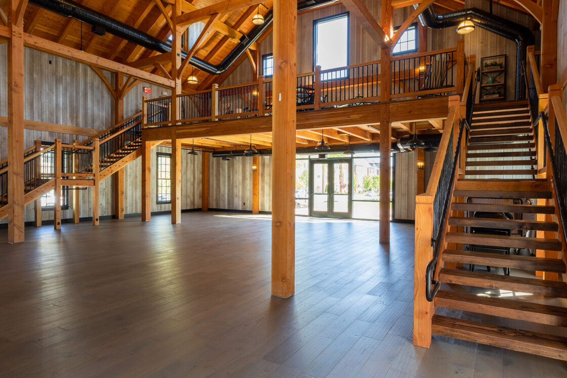 Spacious, rustic interior with exposed wooden beams, high ceilings, large windows, and an open loft area evoke the charm of a bank barn. Warm lighting and wood floors create a welcoming, lodge-like atmosphere.