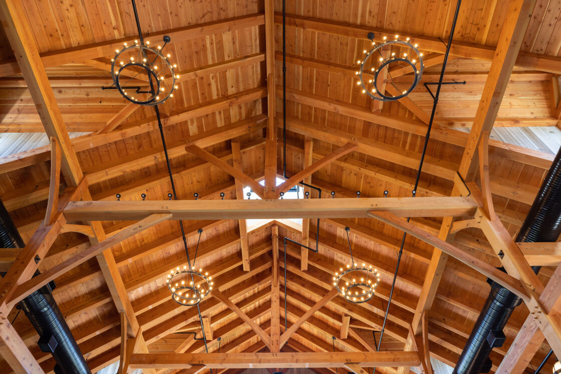 A view of a wooden vaulted ceiling with exposed beams and four wagon-wheel chandeliers, each lit with small bulbs. Black metal air ducts run along both sides, evoking the upscale ambiance of a luxury horse barn or party barn.