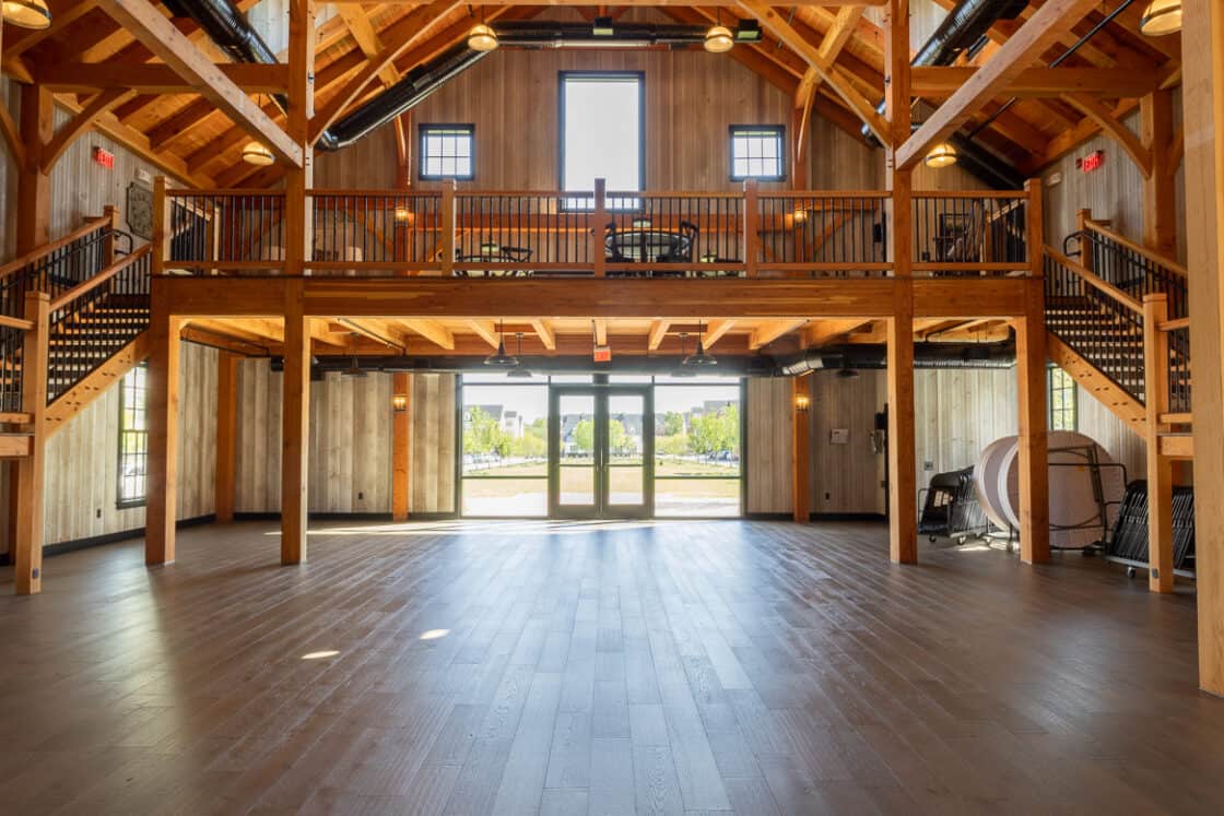 A spacious, rustic party barn interior with exposed wooden beams, high ceilings, chandelier lighting, and a lofted balcony. Large windows and glass doors at the far end let in natural light.