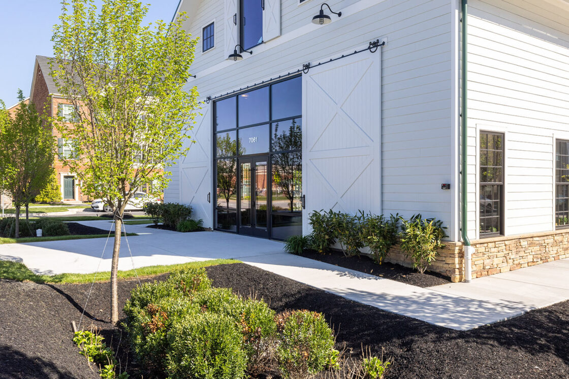 A modern white bank barn–style building with large glass doors, black trim, and a green roof sits among landscaped gardens with shrubs, mulch, and a young tree under a clear blue sky.