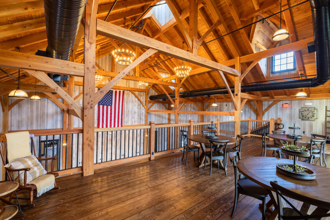 A rustic dining area in a converted bank barn features wooden beams, round tables, black chairs, and an American flag on the wall. Large windows fill the luxury home with natural light, while chandeliers hang from the vaulted ceiling.