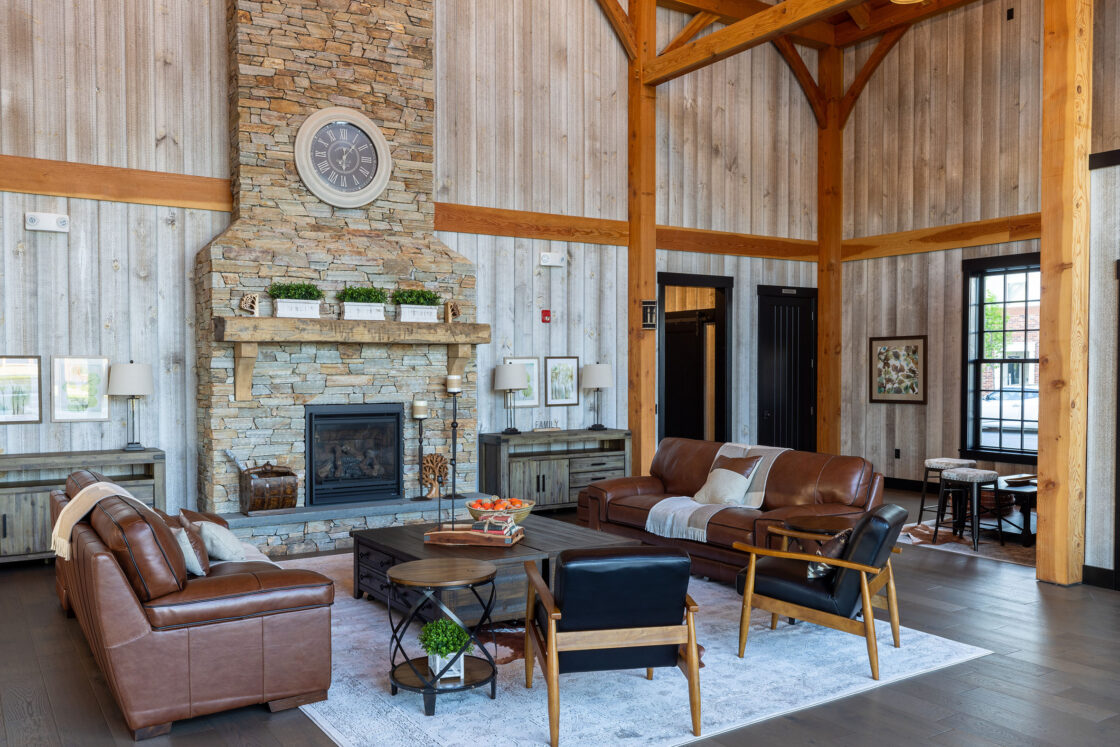 A spacious living room with high wooden beams, stone fireplace, leather sofas, and modern rustic decor evokes the charm of a party barn. Large windows fill this cozy, inviting space with natural light.