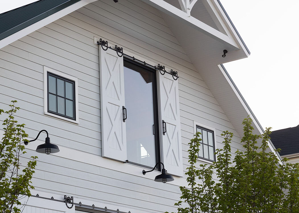 A modern white bank barn-style building with black trim, a large upper window and decorative shutters, smaller side windows, and green trees partially in front. The address 7061 is visible above a glass door.