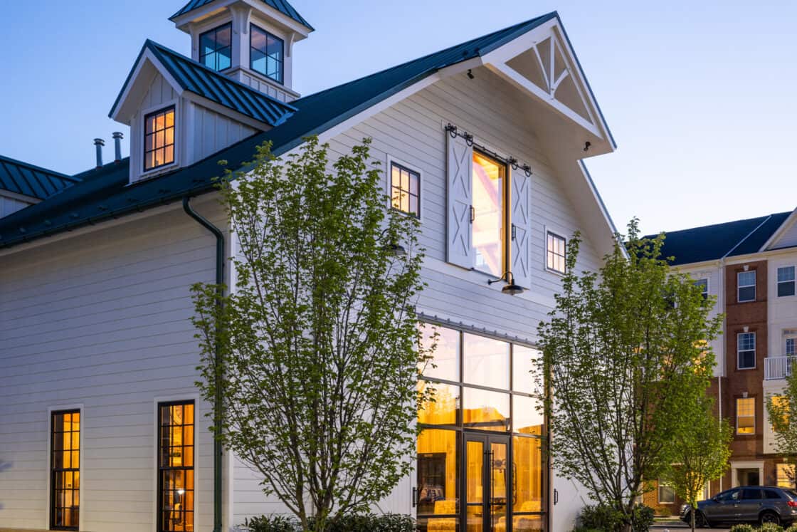 A modern white luxury horse barn with large windows glowing warmly at dusk, surrounded by young trees and neatly landscaped pathways. Nearby, a multi-story brick building is visible.