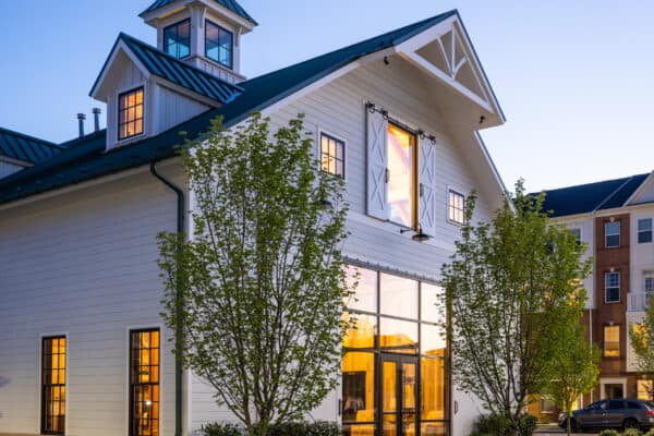 A modern white luxury horse barn with large windows glowing warmly at dusk, surrounded by young trees and neatly landscaped pathways. Nearby, a multi-story brick building is visible.