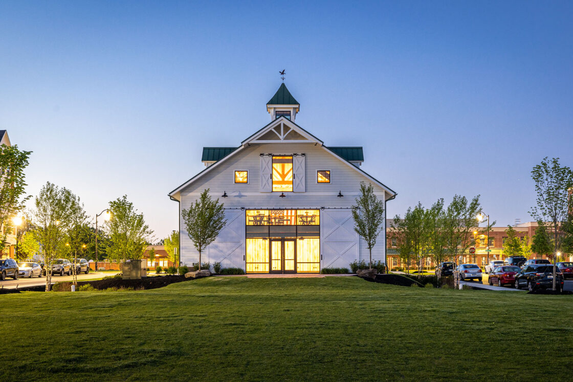 A white bank barn with large windows and a cupola, lit warmly from inside at dusk, stands at the center of a manicured lawn, flanked by trees and parked cars on either side.