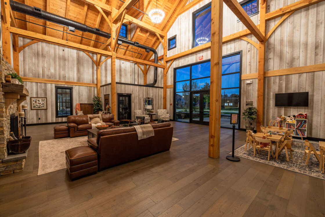 A spacious, rustic-style lounge in a converted bank barn with exposed wooden beams, leather sofas, a stone fireplace, large windows, a TV, and a children’s play area with toys and small furniture on a wooden floor.