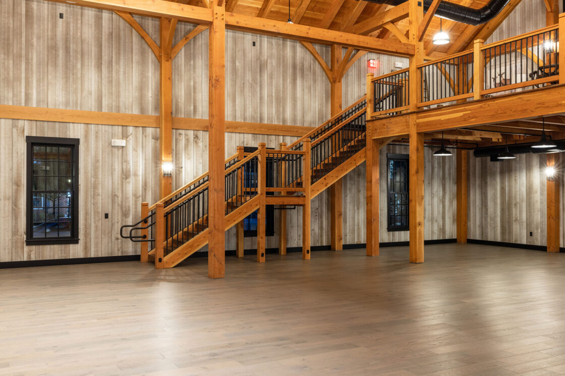 Spacious, empty bank barn with wood floors, exposed beams, and rustic wooden walls. A staircase with black railings leads to an upper balcony. Multiple windows let in natural light, giving the space a warm, inviting feel.