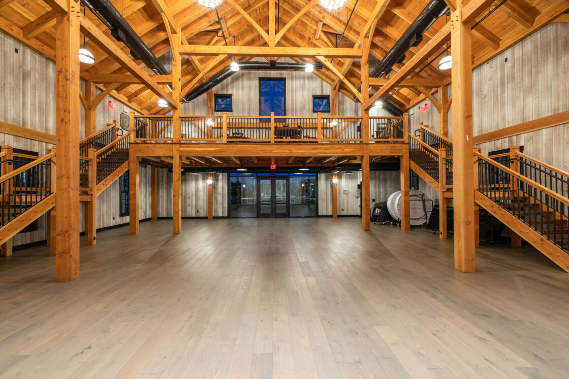 Spacious barn-style hall with wooden beams, high ceiling, two staircases leading to a balcony, metal railings, large windows, and double glass doors at the far end—an ideal party barn in a luxury home with light wood flooring.