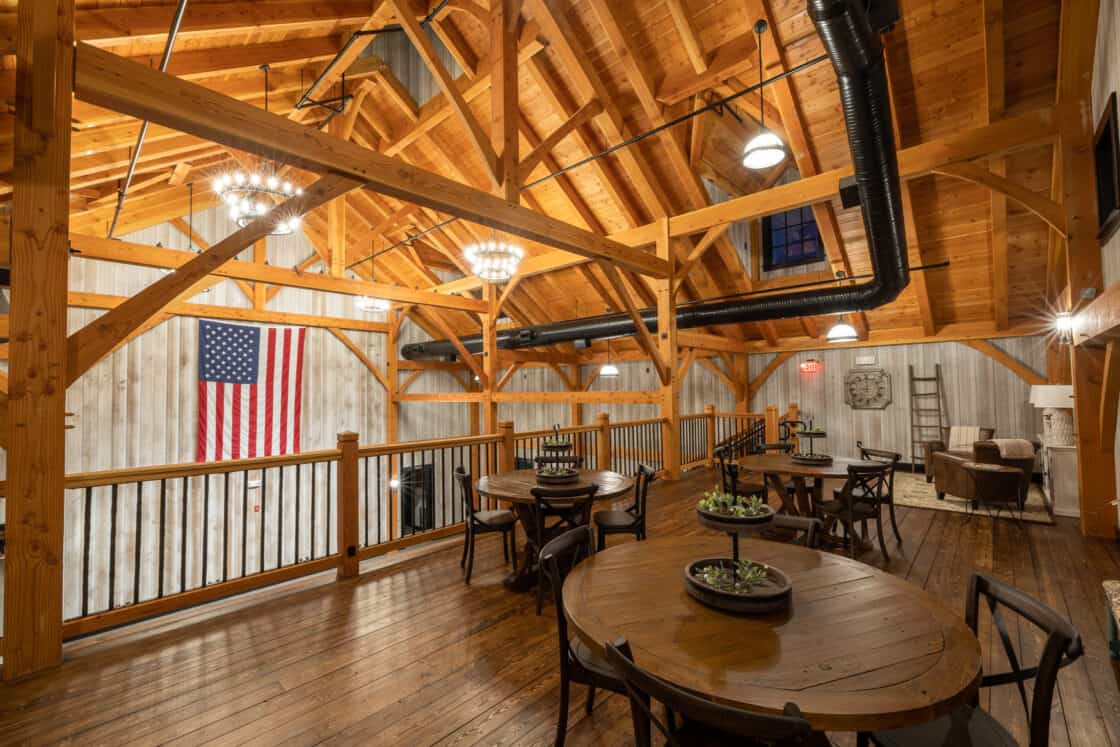Spacious loft with exposed wooden beams, round dining tables, black chairs, chandeliers, and an American flag on a rustic wall—this party barn blends warmth and charm with the inviting feel of a luxury home.