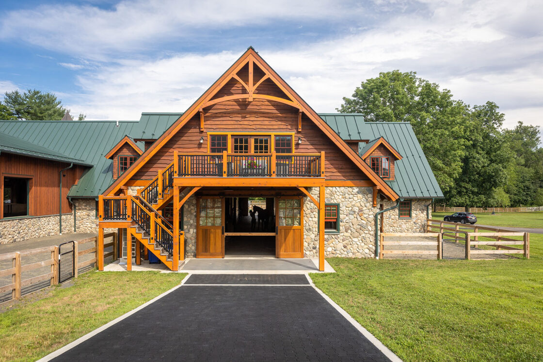 horse barn with visible decorative wooden beams sitting beneath a cloudy sky with black pavement running up to it
