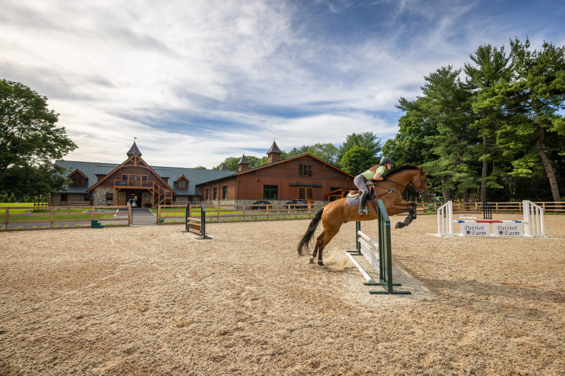 outdoor horse riding area with a horse and rider jumping over a jump which sits beneath a blue cloudy sky