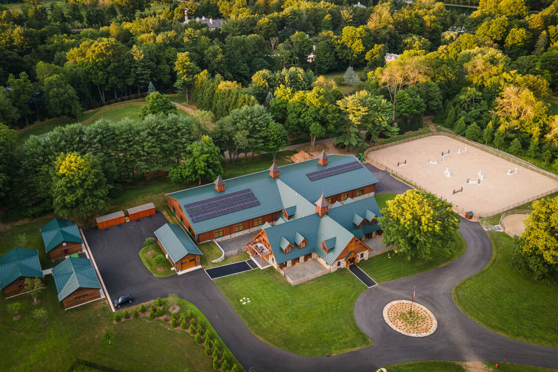 drone view of a custom horse barn with a green roof sitting in front of green trees with an outdoor riding area to its right