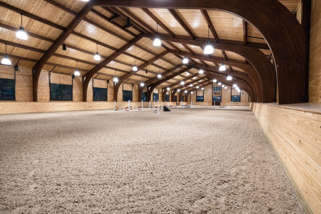 indoor riding arena that is completely empty sitting beneath illuminated light fixtures and wooden ceiling beams