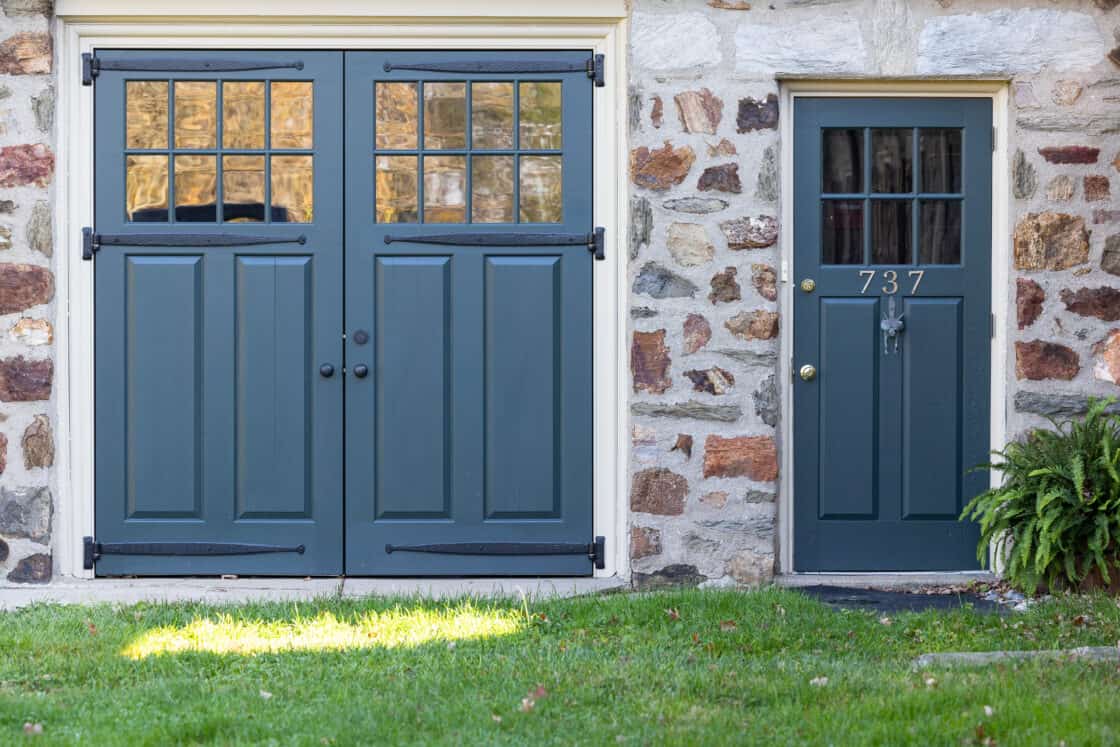 Blue double barn-style doors and a matching single door with the number 737 grace this stone bank barn; grass fills the foreground, and a fern plant sits beside the door, blending rustic charm with luxury home appeal.