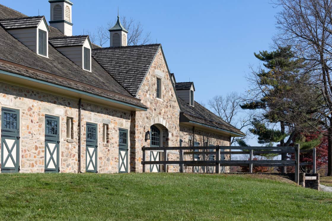 A stone bank barn with green doors and cupolas sits on a grassy lawn under a clear blue sky, bordered by a wooden fence and nearby trees.
