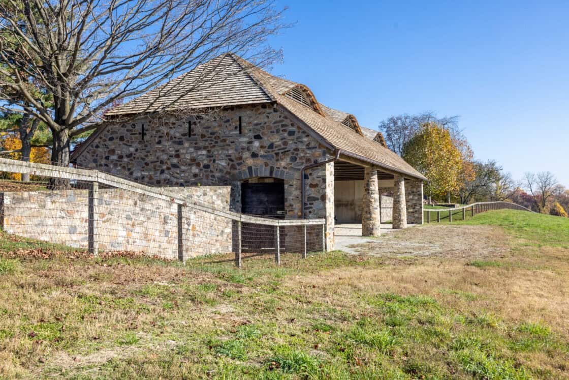 A rustic stone bank barn with a wooden shingle roof stands behind a stone and wire fence, surrounded by grass and autumn trees under a clear blue sky.