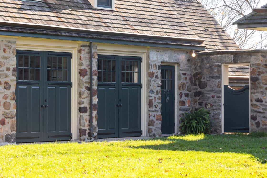Stone building with wooden double doors and a single door, all painted dark green. Reminiscent of a classic bank barn, a small fern sits by the single door while sunlight highlights the grass lawn in front.