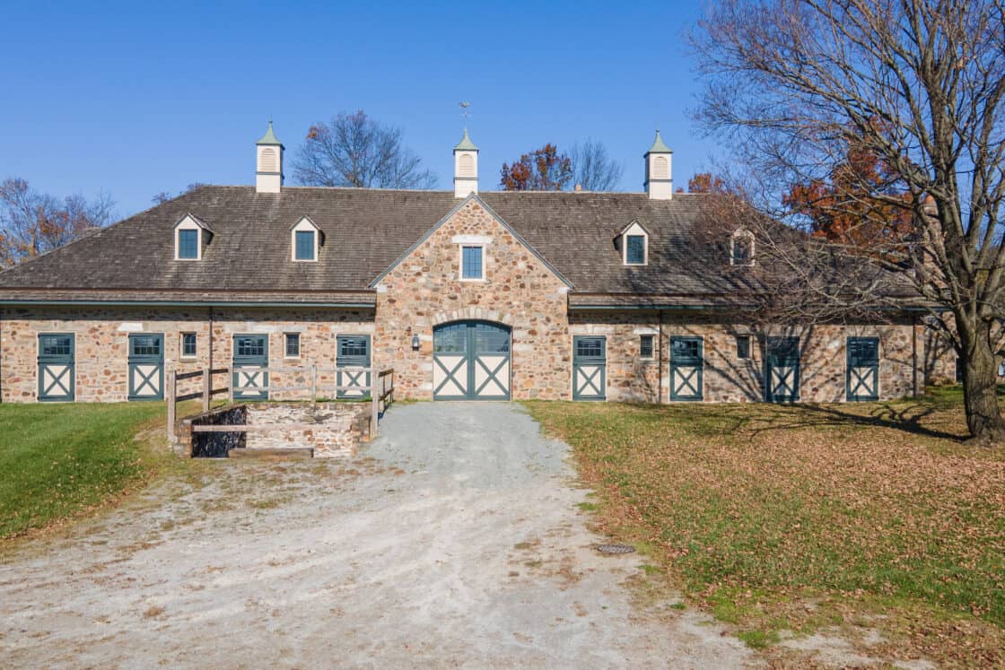 A large brick bank barn with green and white doors and cupolas stands under a clear blue sky. Leafless trees and scattered autumn leaves surround the building, with a gravel path leading to the entrance.