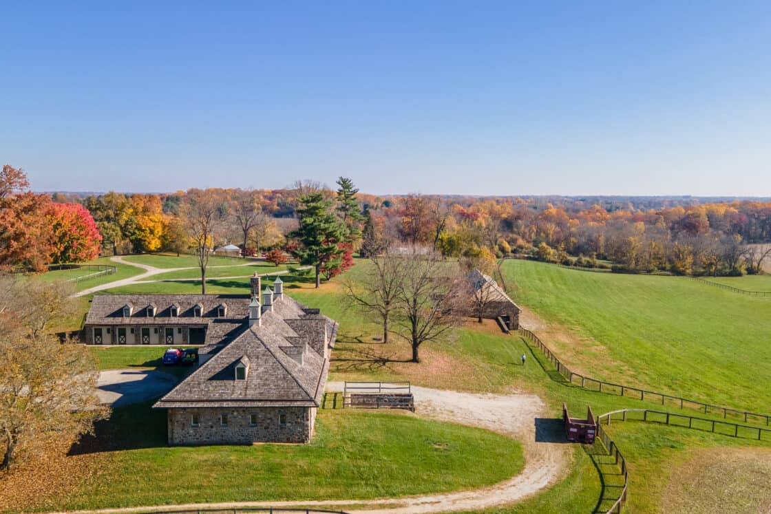 Aerial view of a rustic stone bank barn with a wooden roof, surrounded by green fields and trees with autumn foliage under a clear blue sky. A dirt road curves around the property.