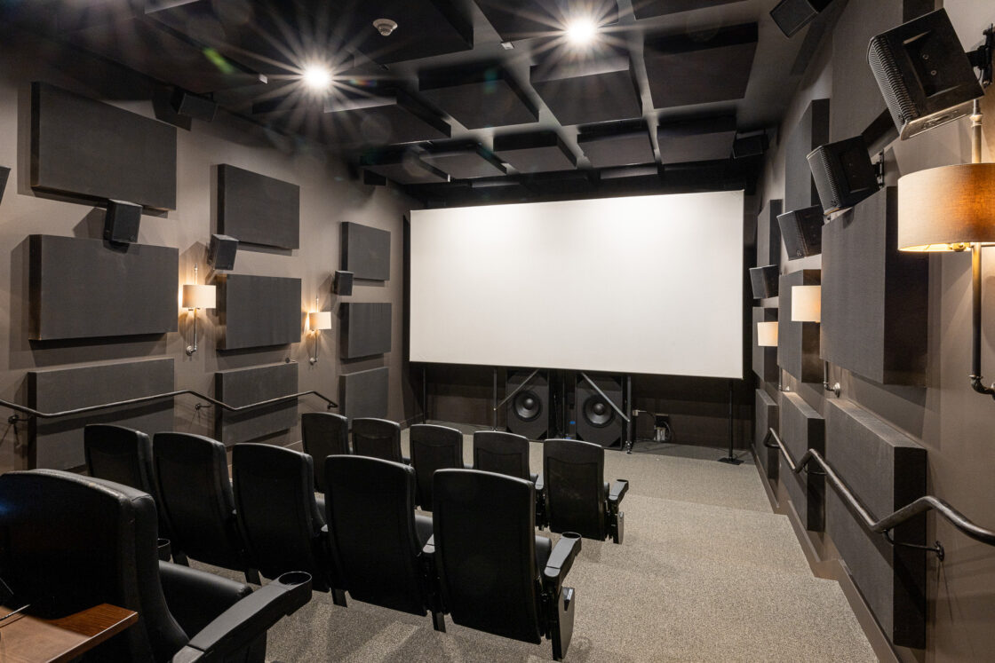 A modern luxury home theater room with black cushioned seats in rows, a large white projection screen, wall-mounted speakers, soundproof panels, and soft wall lighting—perfect for bringing a party barn vibe indoors.