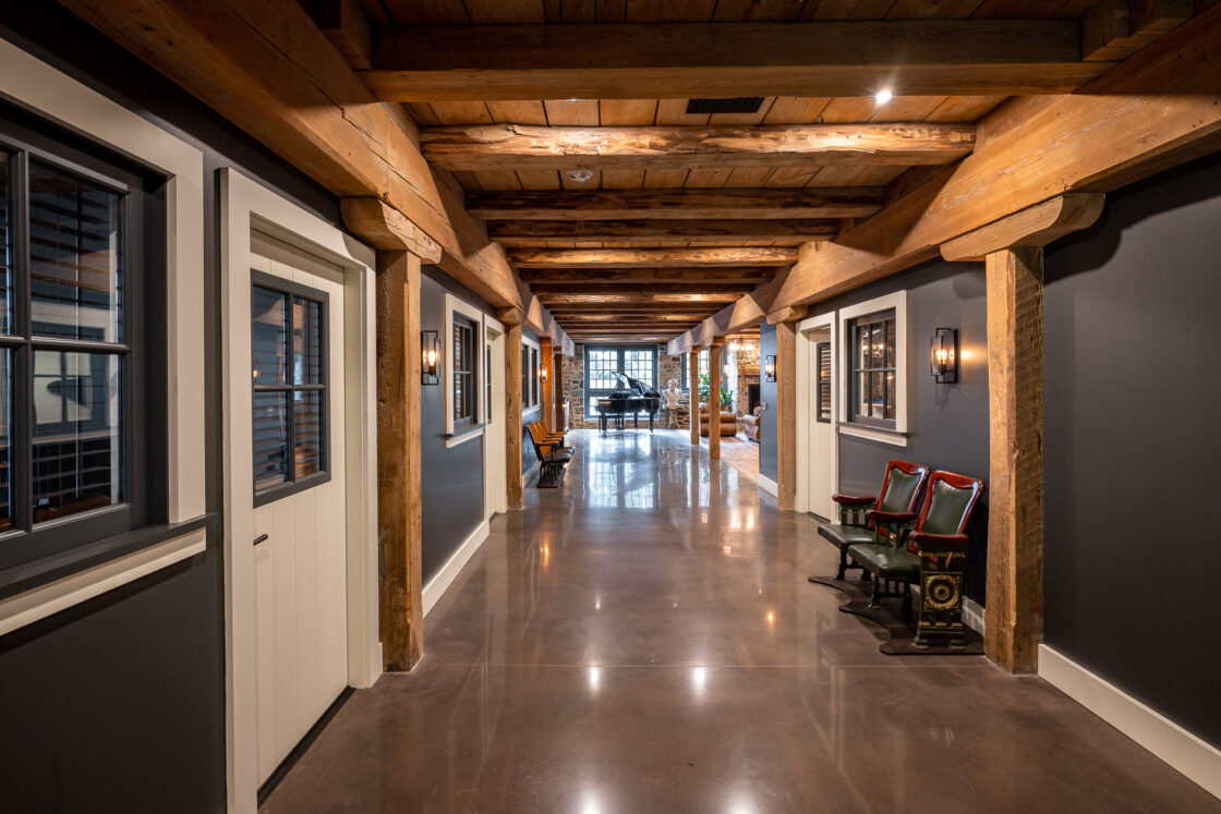 A spacious hallway with exposed wooden beams, polished concrete floors, gray walls, and vintage chairs evokes the charm of a luxury home inspired by a restored bank barn, with a distant view of a piano in the well-lit rustic-modern interior.