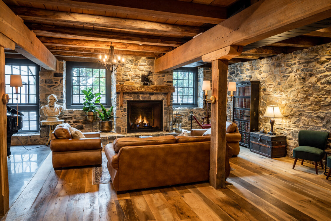 A rustic living room in a luxury home with exposed wooden beams, stone walls, leather sofas, a fireplace, large windows, indoor plants, a bust sculpture, bookshelves, and warm, ambient lighting.