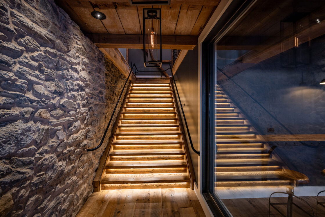 A wooden staircase with built-in step lighting ascends between a rustic stone wall and a glass panel, evoking the charm of a luxury horse barn while creating a warm and inviting ambiance.