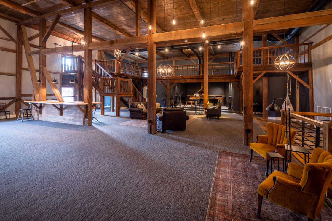 Spacious, rustic interior with exposed wooden beams and a loft area, this party barn features modern lighting, cozy armchairs, sofas, and a carpeted floor—blending vintage and contemporary styles in an open lounge setting.