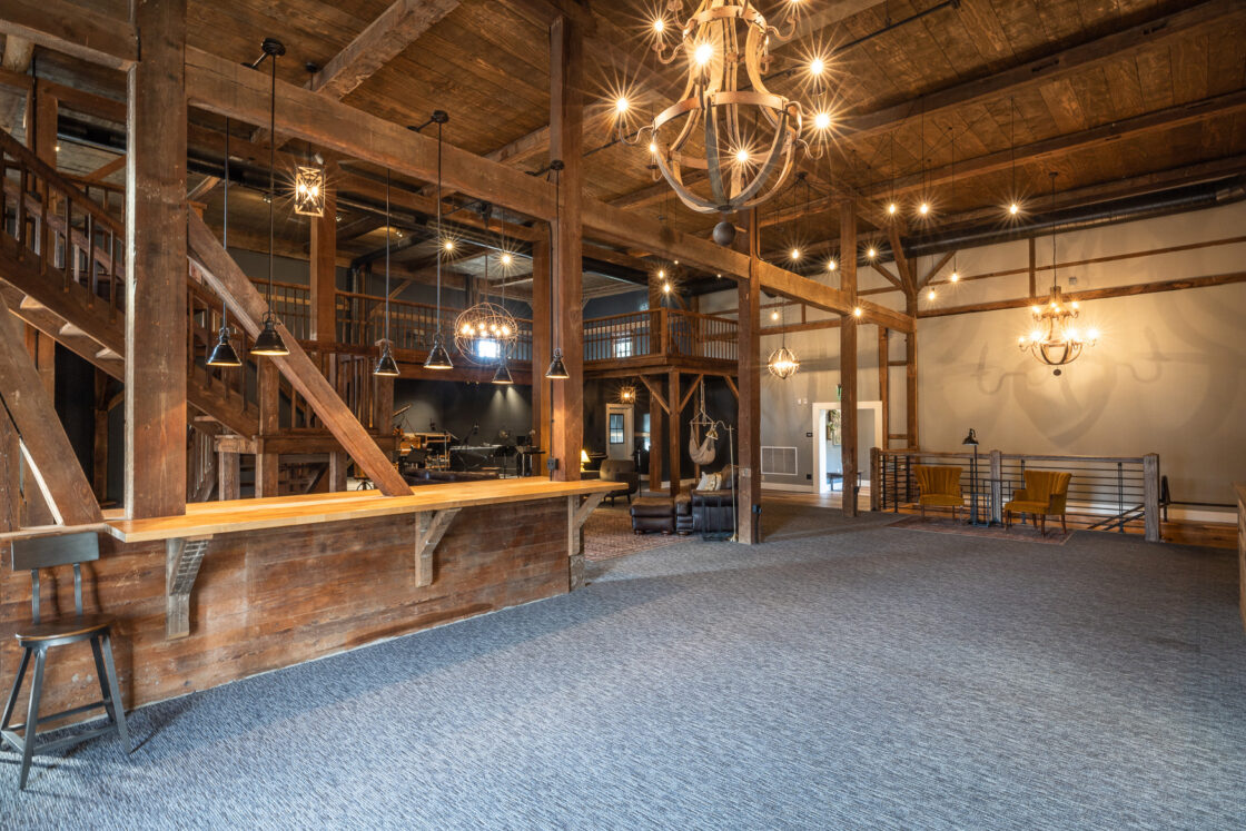 A spacious, rustic interior reminiscent of a luxury horse barn, with exposed wooden beams, chandeliers, a wooden bar with stools, carpeted floors, lounge furniture, and stairs to a mezzanine. Warm lighting adds cozy ambiance.