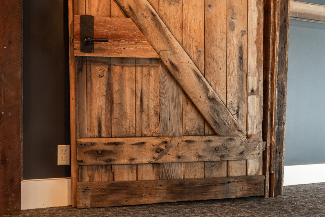 A rustic wooden door with a diagonal support beam, black metal handle, and latch stands partly open against a dark wall and grey carpeted floor—perfect for a luxury horse barn or party barn setting.