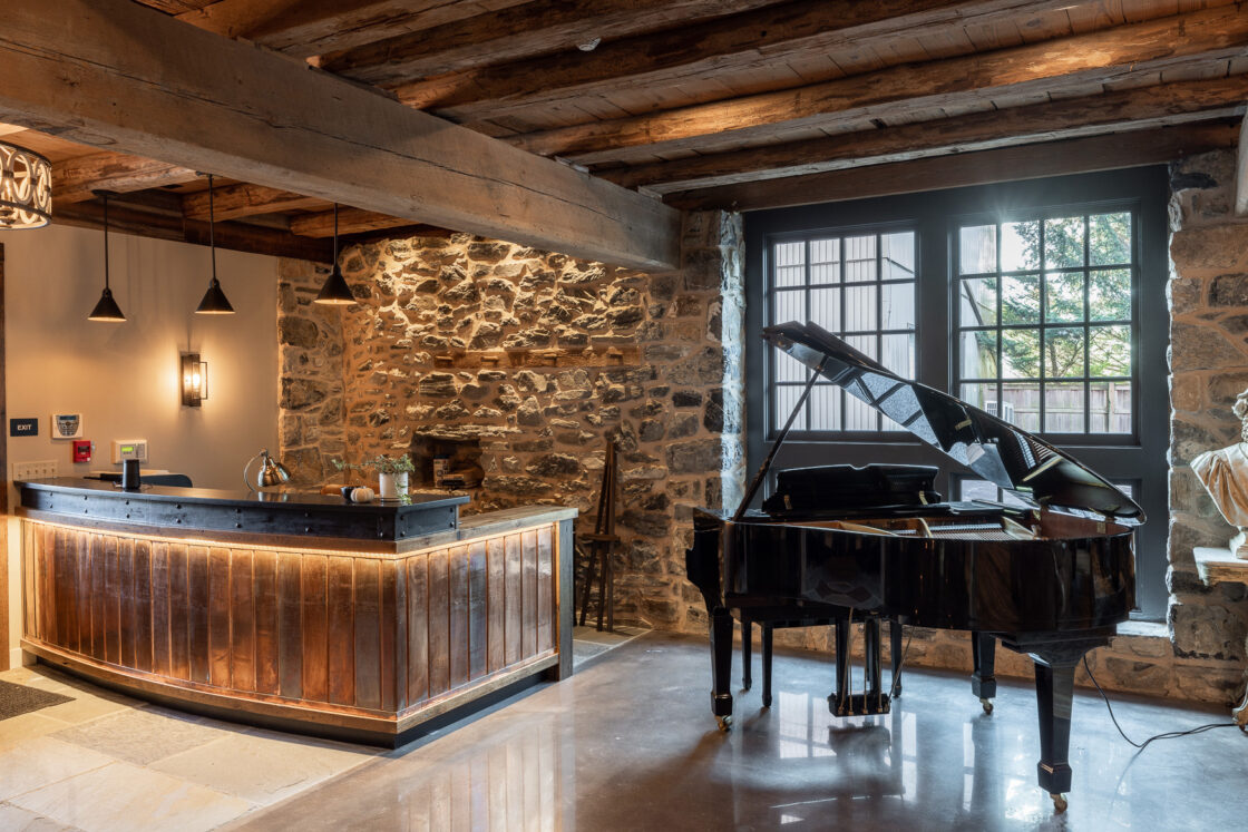 A cozy room in a luxury horse barn with exposed stone walls and wooden beams features a grand piano on a polished floor and a curved wooden reception desk with pendant lights overhead. Large windows let in natural light.