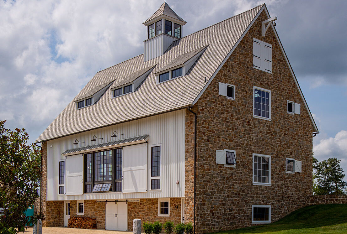 A large bank barn with stone and white siding sits on a grassy hill under a partly cloudy sky. The barn has multiple windows, a cupola on the roof, and a gravel driveway in front.