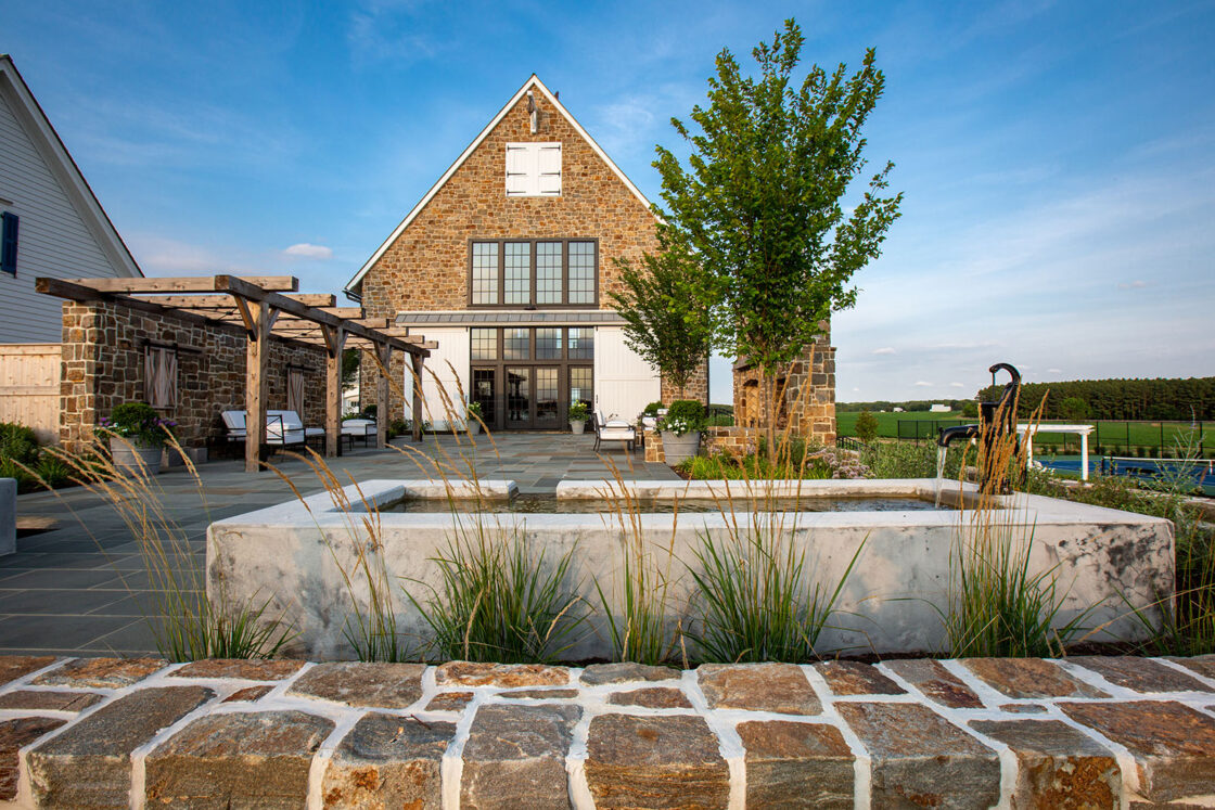A modern farmhouse with stone walls, large windows, and a gabled roof stands behind a landscaped patio featuring seating areas, a pergola, ornamental grasses, and a low stone wall—perfectly complementing the luxury home’s elegant style.